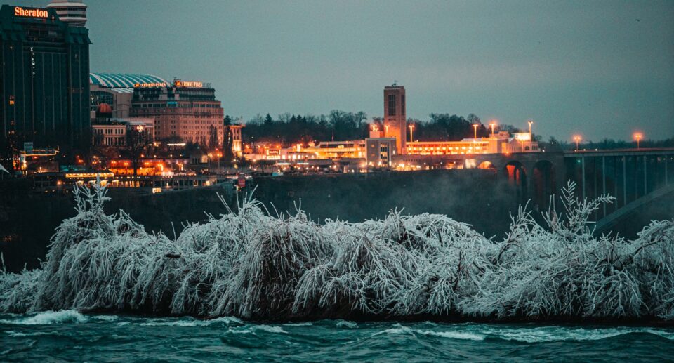 Niagarafälle im Winter mit vereisten Bäumen und beleuchteter Stadt-Skyline in der Dämmerung